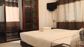 A hotel room with a bed, a television mounted on the wall, and a sliding glass door leading to a balcony. - Sai Pavilion (Puttaparti)