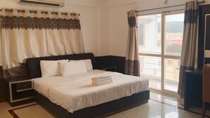 A hotel room with a bed, a desk, a chair, a window with curtains, and a view of the outside. - Sai Pavilion (Puttaparti)
