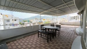 A covered outdoor area with a table and chairs, a patterned floor, and a view of buildings in the distance. - Sai Pavilion (Puttaparti)