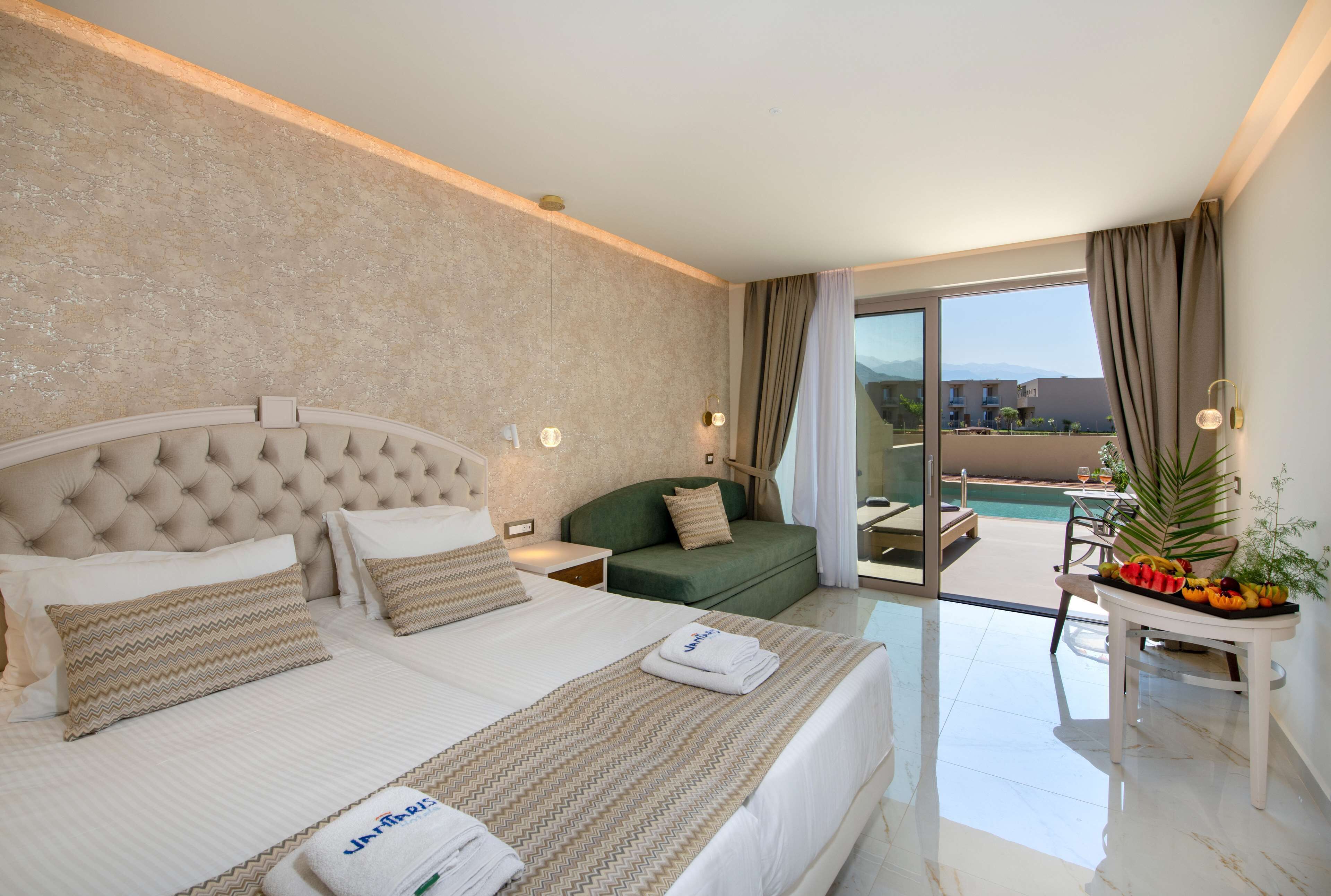 A hotel room with a large bed, a green sofa, a small table with fruit, and a balcony with a pool.