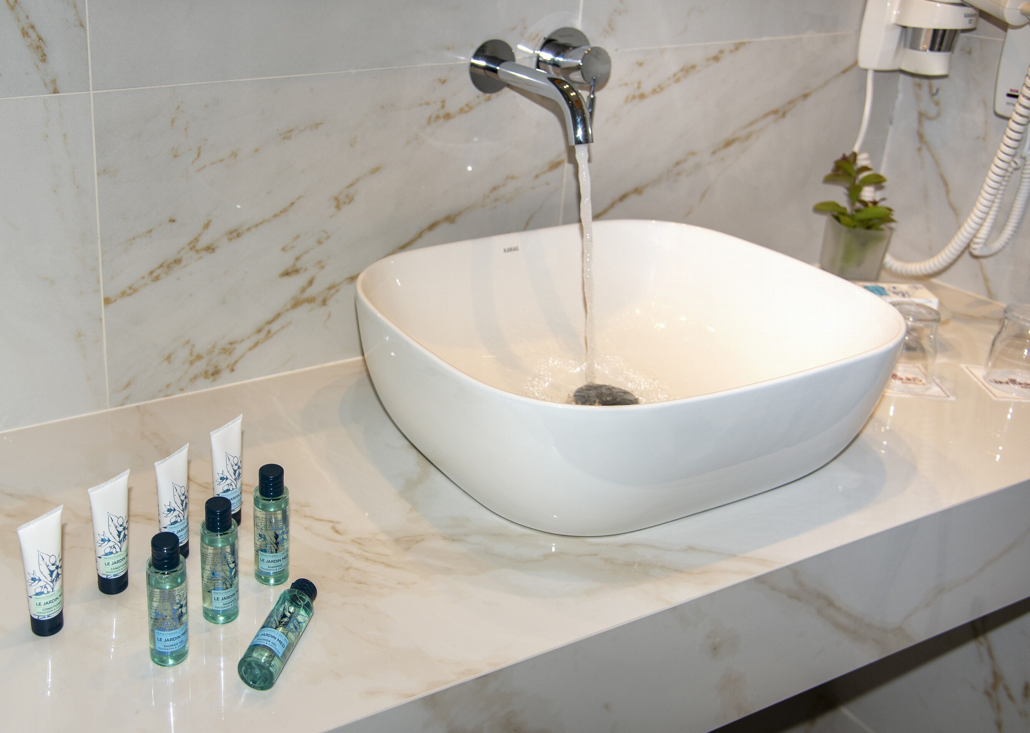 Une salle de bain avec un lavabo en marbre blanc, de l’eau courante et plusieurs flacons de produits de toilette posés sur le comptoir.