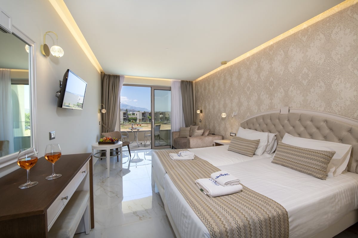 Quarto de hotel com uma cama grande, uma mesa com dois copos de vinho, uma televisão e uma varanda com mesa e cadeiras.