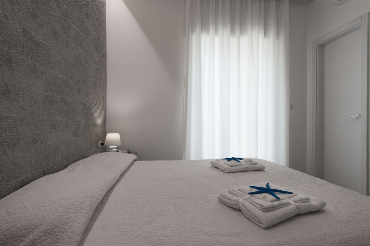 Una habitación de hotel moderna con una cama grande, una mesita de noche con lámpara y una ventana con cortinas traslúcidas.