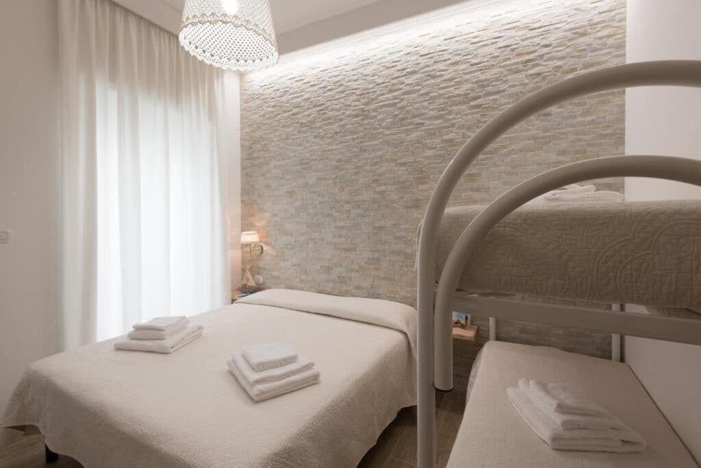 Quarto de hotel com beliche, cama arrumada com toalhas, parede com textura de pedra e luminária suspensa.