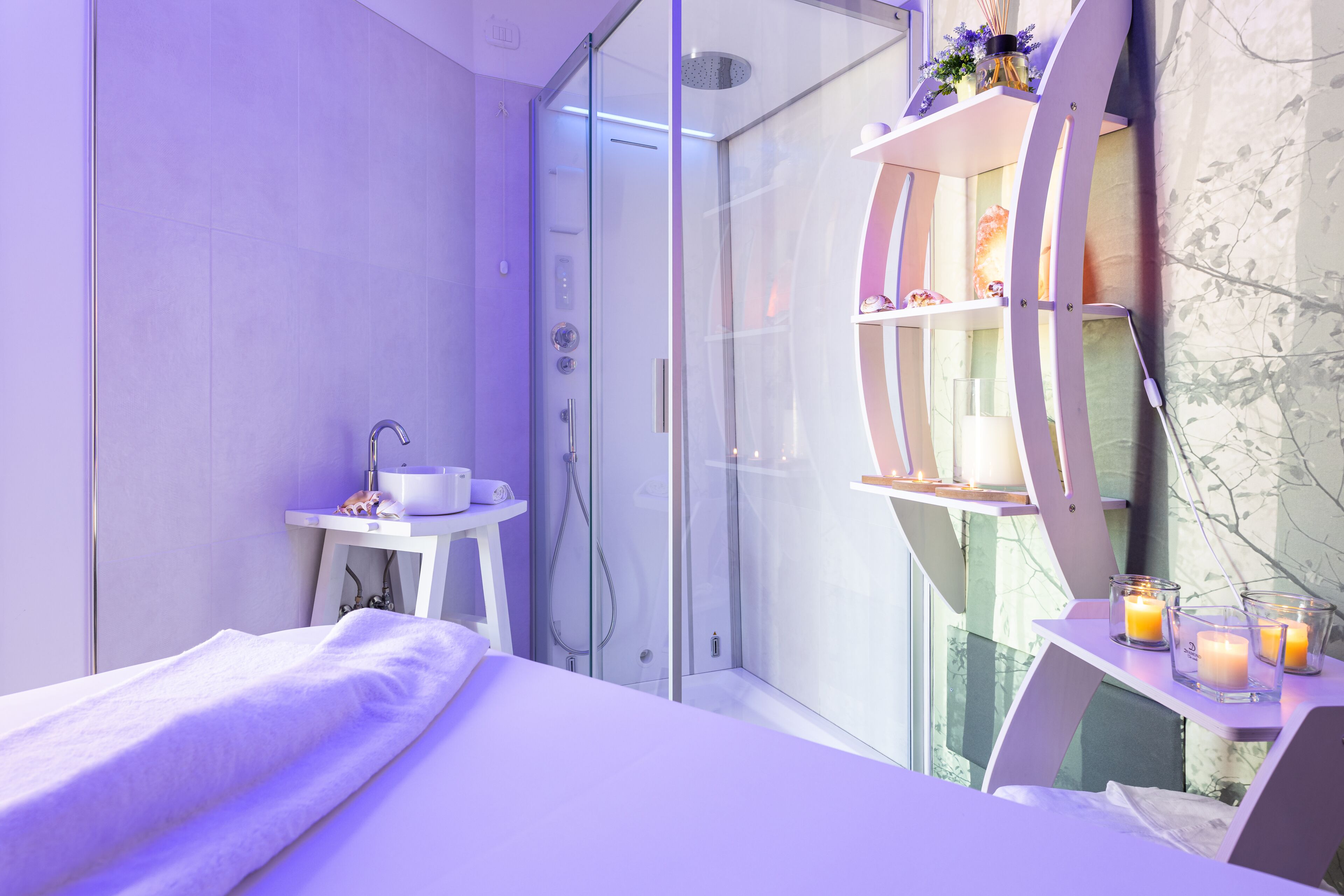 A modern bedroom with a glass shower, a bedside table with candles, and a white bed.