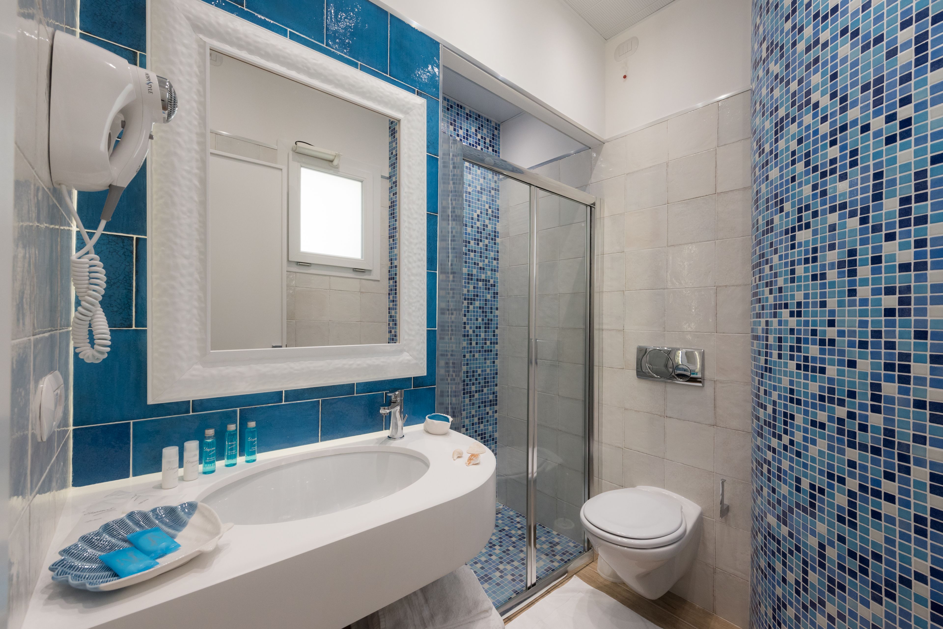 Une salle de bain avec un lavabo blanc, des murs recouverts de carreaux bleus, une douche en verre et des toilettes blanches.