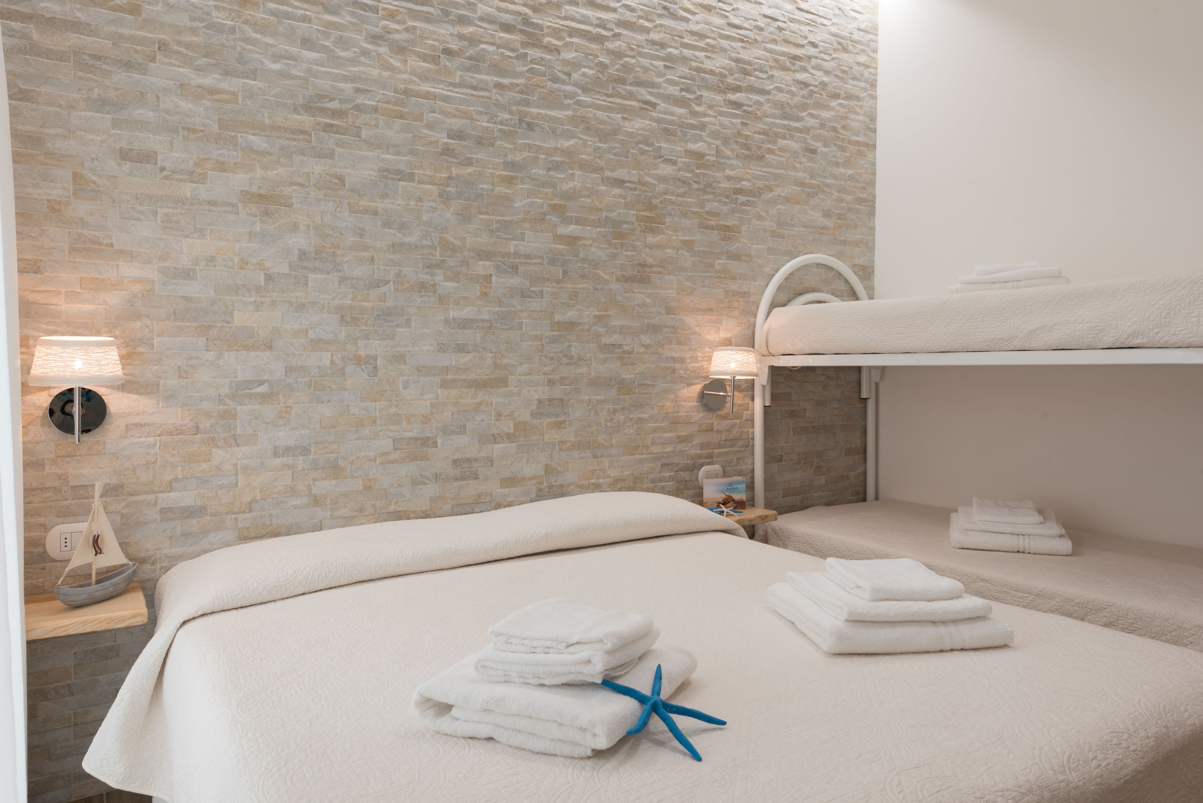 Ein Zimmer mit Steinwand, einem Bett mit weißen Leinen, einem Nachttisch mit Lampe und einem kleinen Regal mit einem dekorativen Gegenstand.