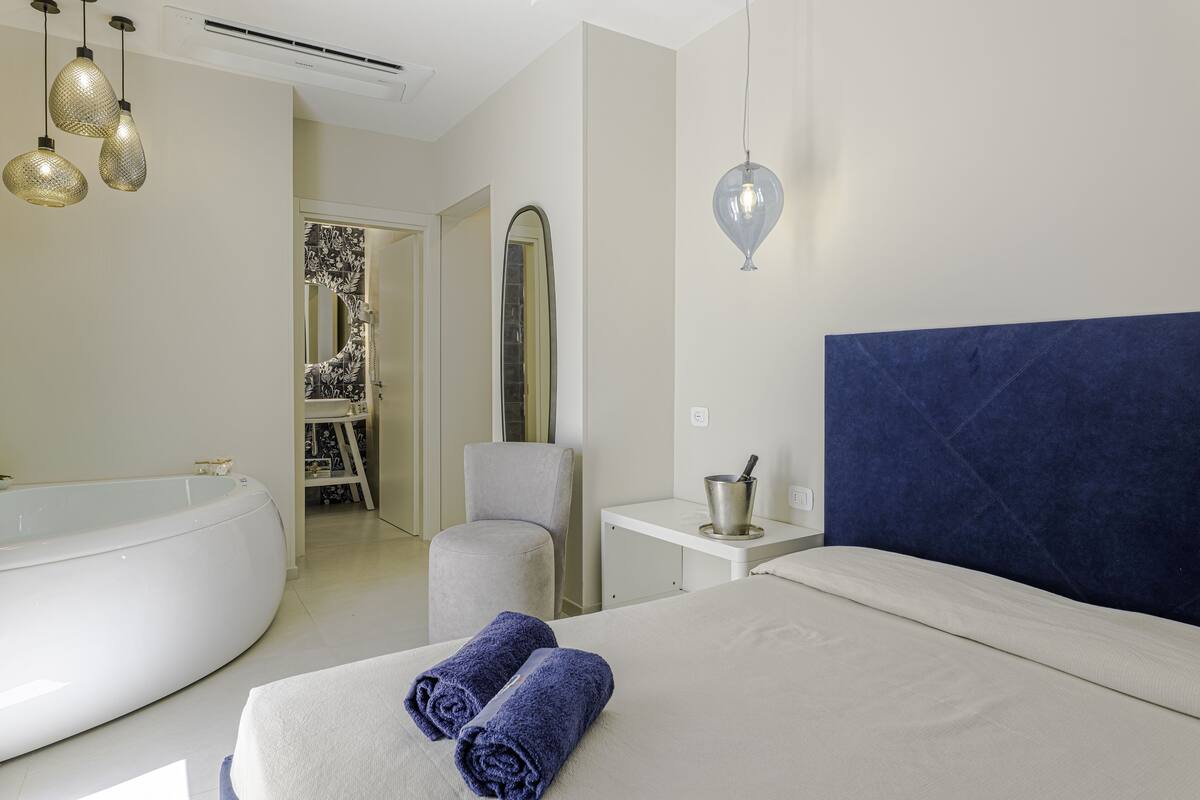 Una habitación de hotel moderna con una bañera grande, una cama con cabecera azul y una mesita de noche pequeña con una lámpara.