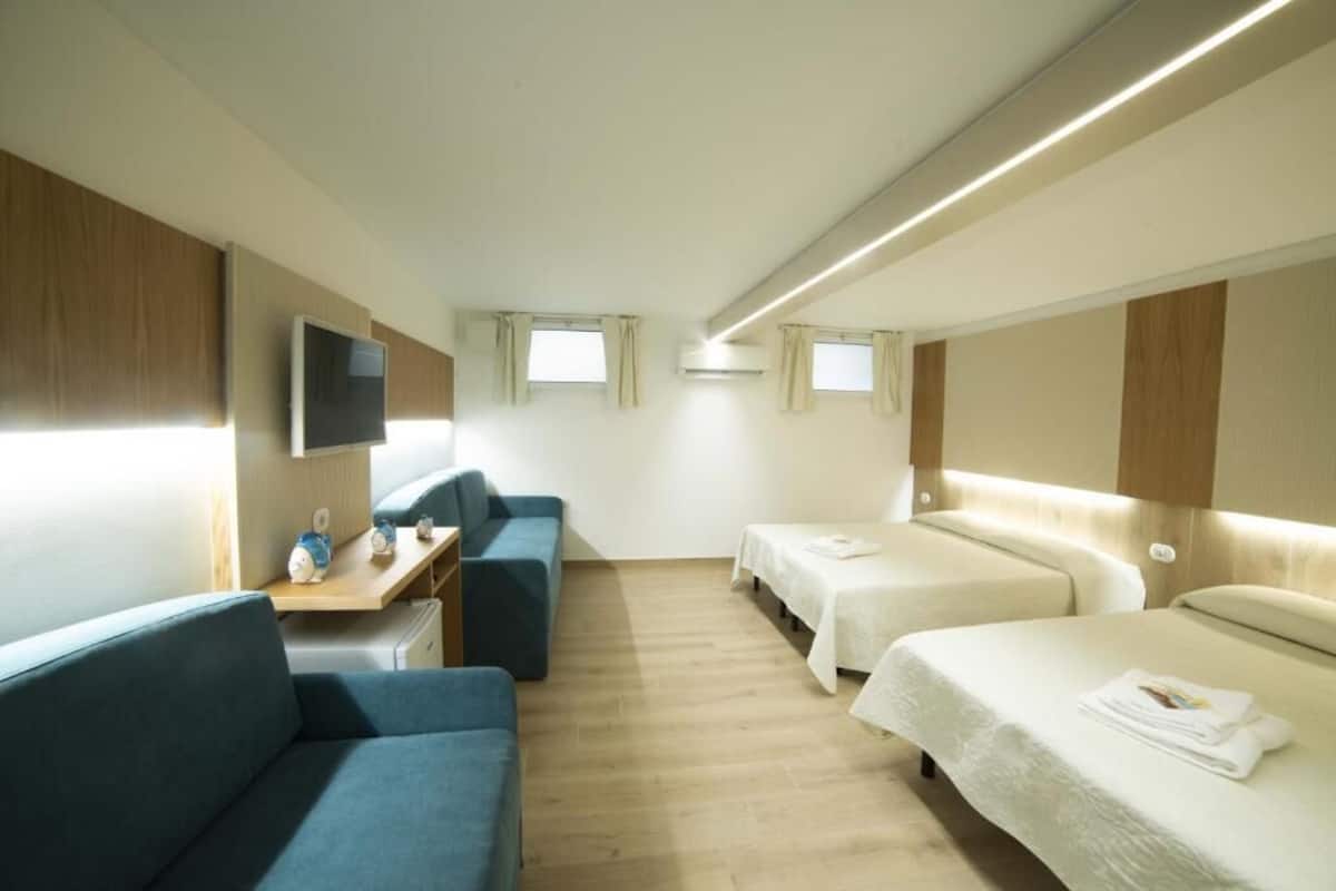Una habitación de hotel compacta con sofá, dos camas individuales, una mesita con lámpara y televisor instalado en la pared.