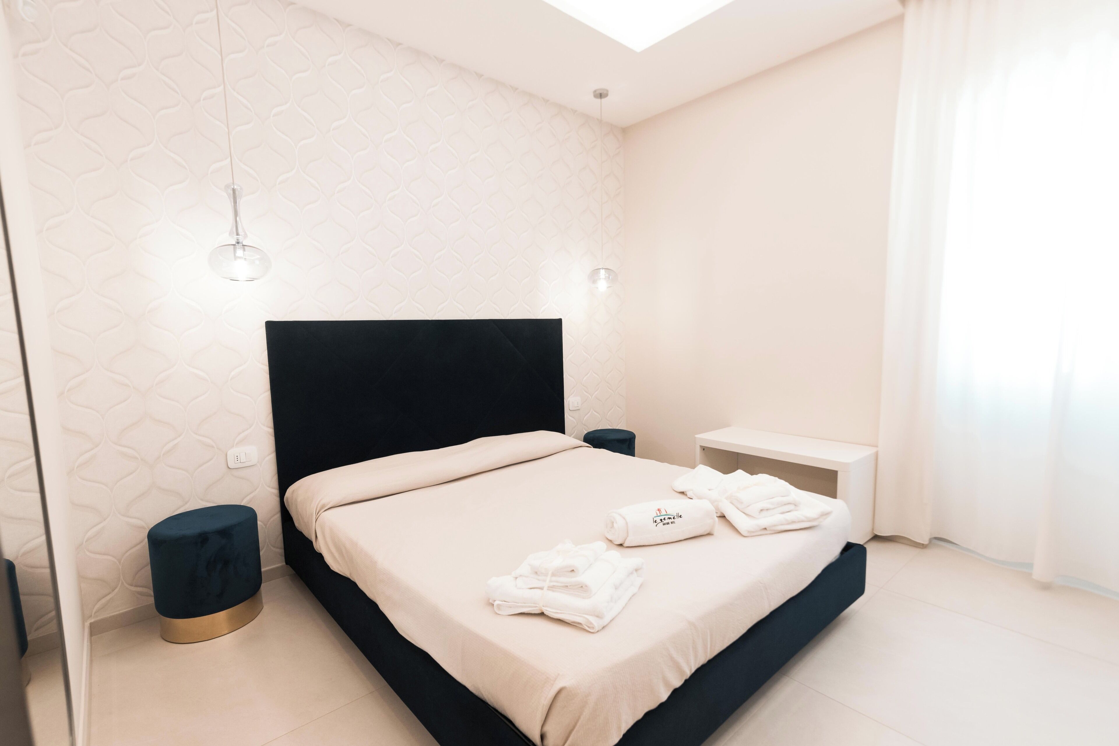Un dormitorio moderno con cabecera negra, colcha color beige, dos mesitas de noche y un taburete pequeño.