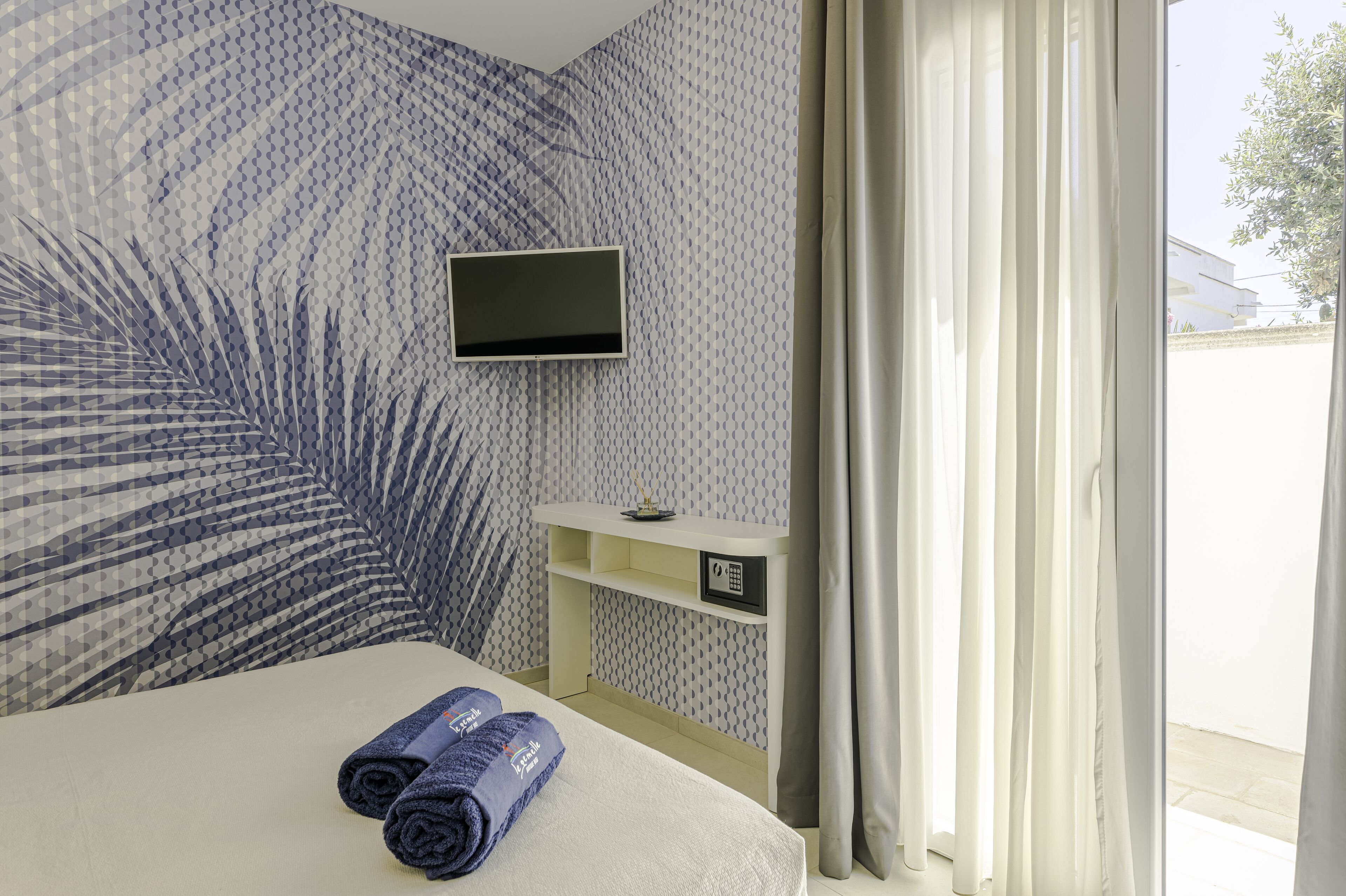 Een hotelkamer met een bed, een aan de muur bevestigde televisie, een klein bureau met een kluisje en een raam met gordijnen.