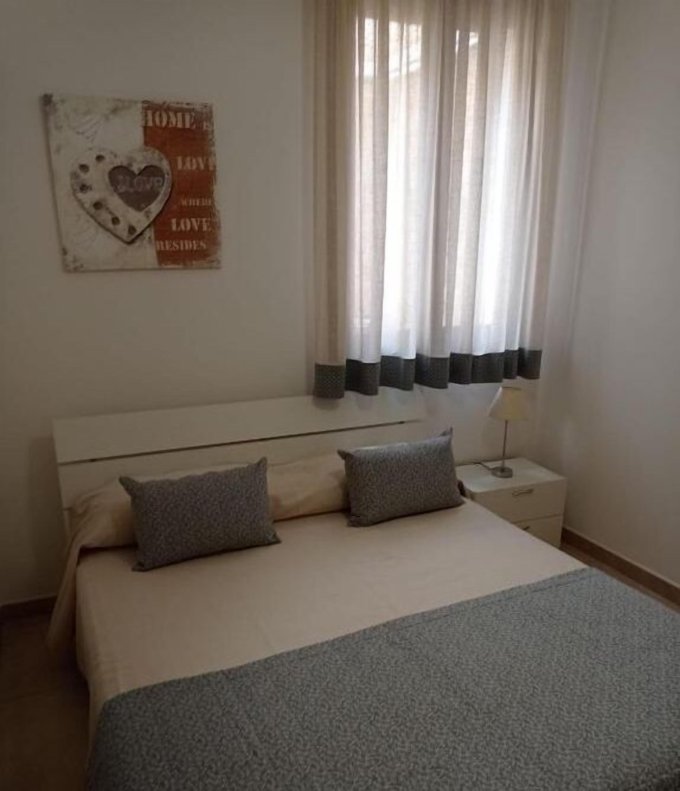 A bedroom with a bed, two pillows, a nightstand with a lamp, a wall decoration, and a window with sheer curtains.