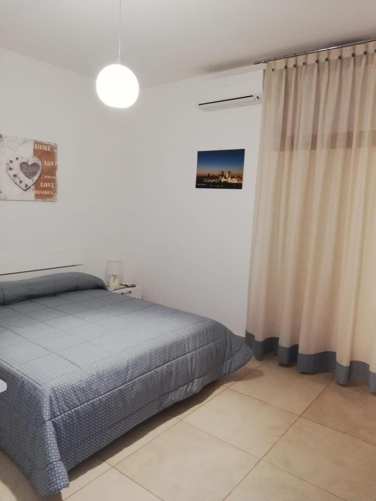 A bedroom with a bed, a nightstand, a wall-mounted air conditioner, a ceiling light, a window with sheer curtains, and a framed picture on the wall.