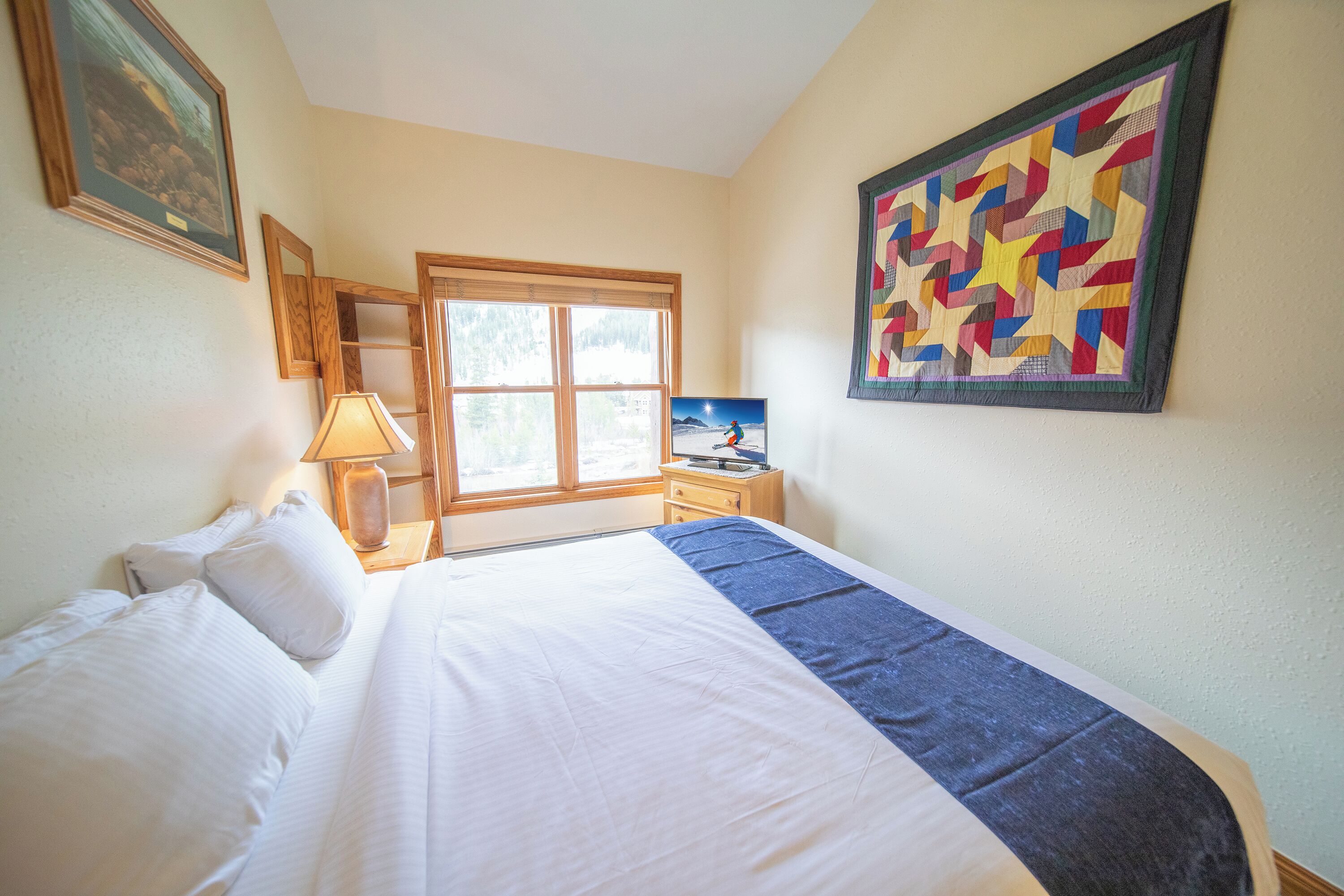A neatly made bed with white linens, a bedside lamp, a wooden nightstand, and a window with a view of trees.