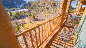 Un balcón de madera con sillas de metal, con vista a un paisaje montañoso que incluye una cabaña.