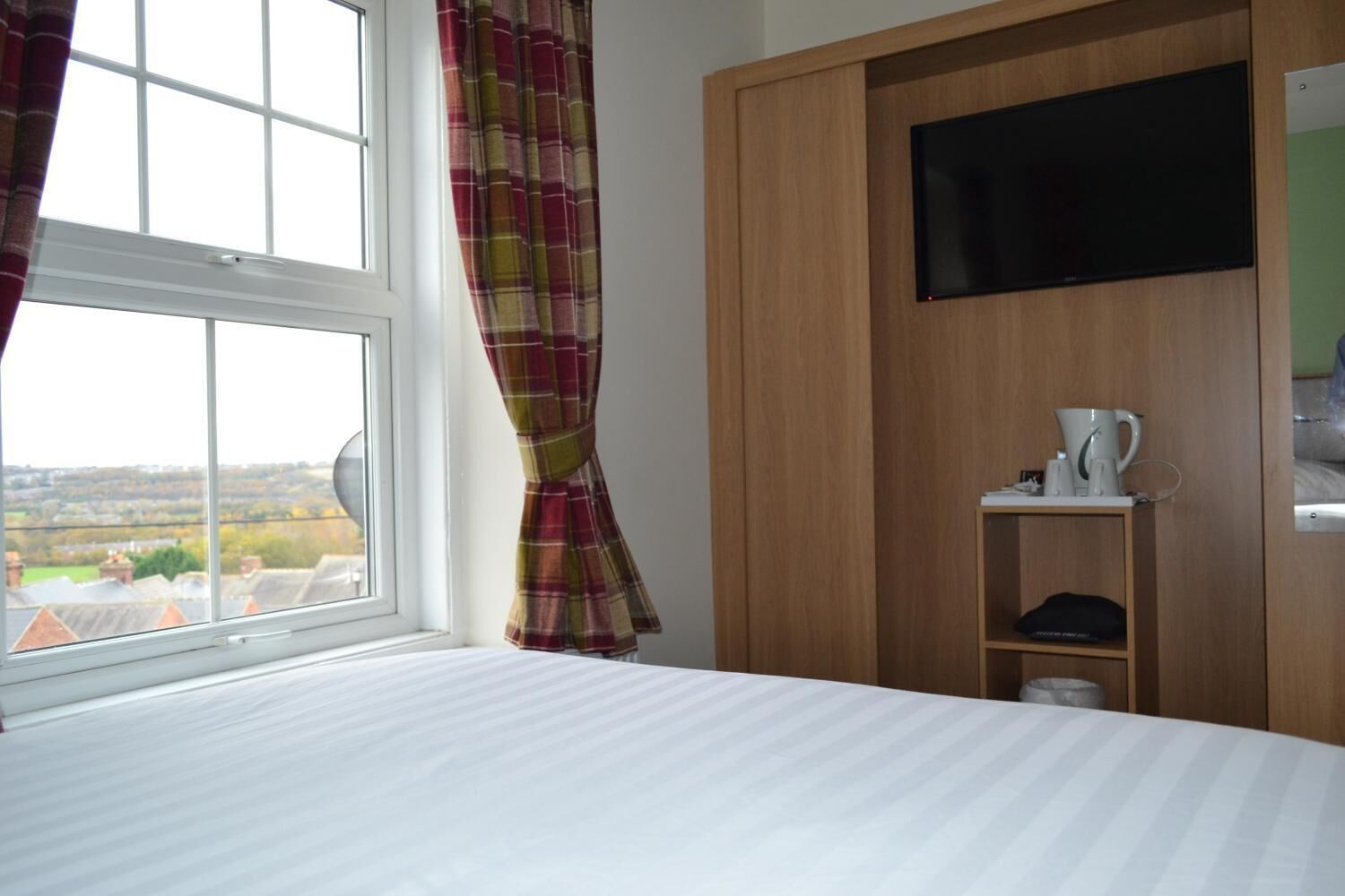 Habitación de hotel con cama, televisor montado en un mueble de madera, ventana con cortinas y vistas al exterior.