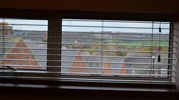 Una ventana con persianas horizontales, vista a un edificio de ladrillo con tejados puntiagudos y un paisaje verde en la distancia.