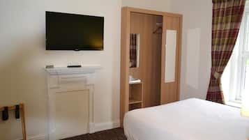 A hotel room with a flat-screen TV mounted on the wall, a wooden wardrobe, a white bed, and a window with plaid curtains.