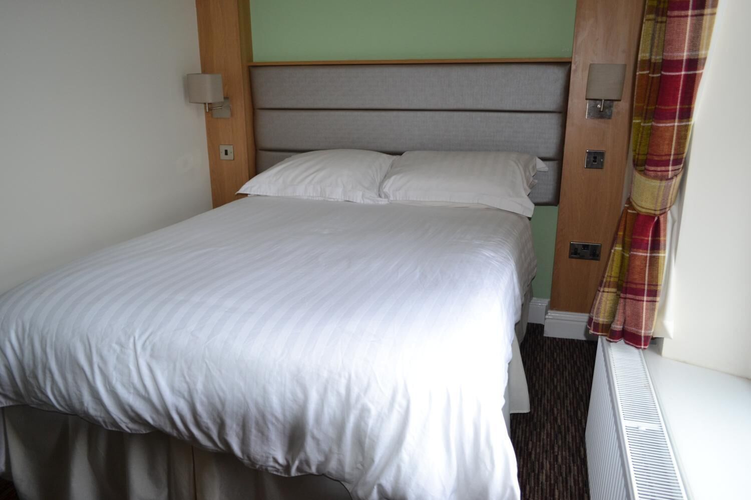 Una cama individual con cabecera gris, ropa de cama blanca y un aplique fijado a la pared.