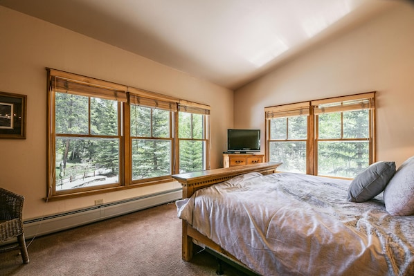 A bedroom with large windows, a bed, a TV, and a wooden nightstand.