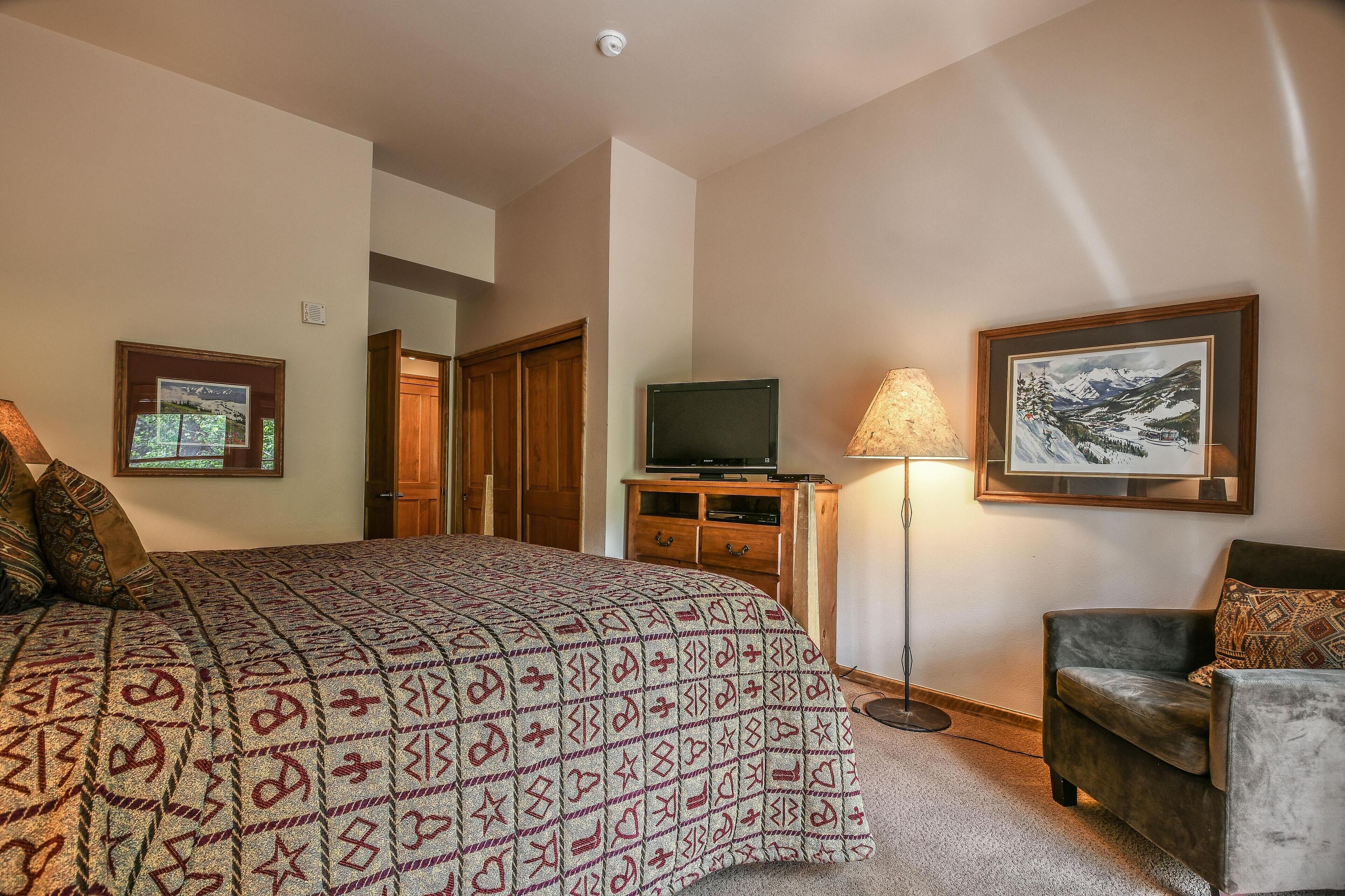 Ein Schlafzimmer mit einem großen Bett, einem Fernseher, einer Lampe, einem Sessel und gerahmten Bildern an der Wand.