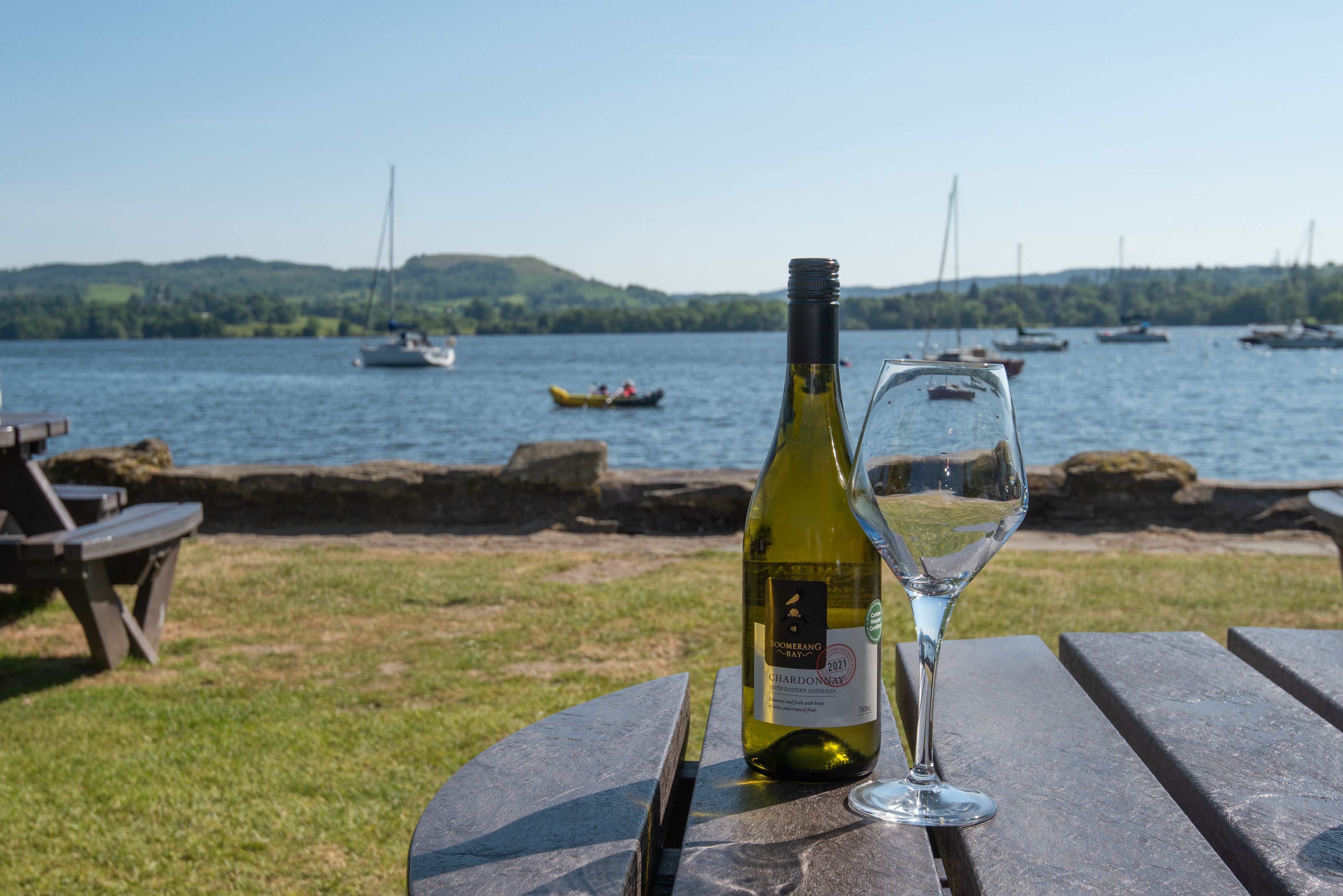 A bottle of wine and a glass on a wooden table by a lakeside with boats in the background.