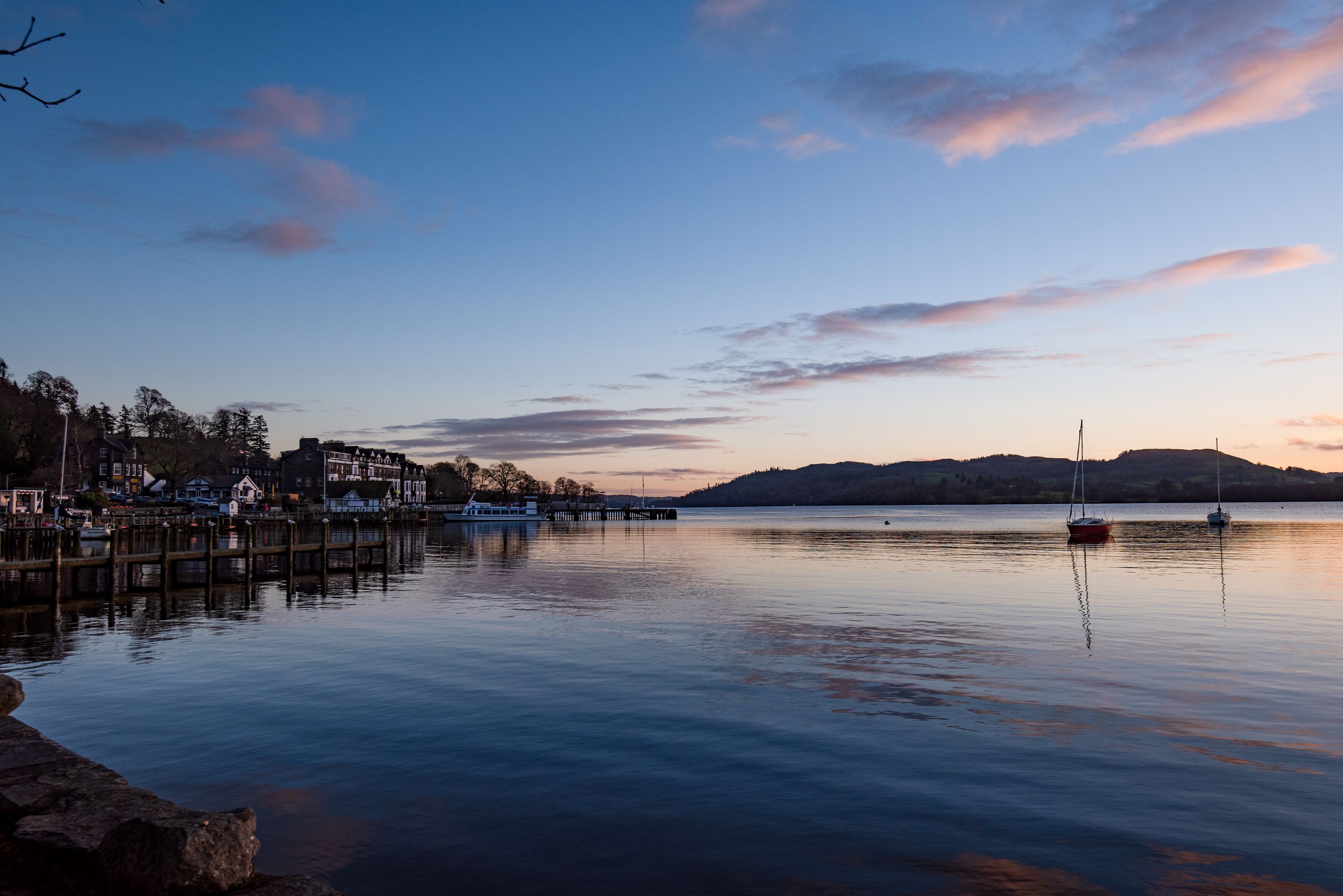 A calm lake with a dock and boats, surrounded by buildings and hills in the background.