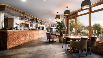 Uma área de bar com balcão em padrão herringbone, várias torneiras e uma televisão na parede. Há várias mesas e cadeiras dispostas para acomodar os clientes, e o espaço é iluminado por luminárias pendentes.
