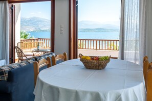 A dining area with a table set for two, a wicker fruit basket, and a view of a lake and mountains through open doors.