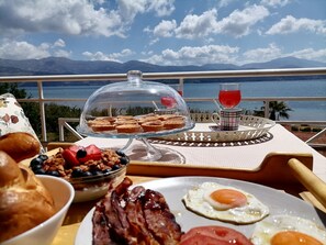 A breakfast table with pastries, a glass of red drink, and a scenic view of mountains and water.