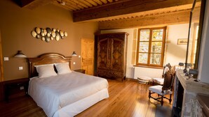 Un dormitorio con una cama grande, suelo de madera, un armario, una chimenea y una ventana con vistas a árboles.