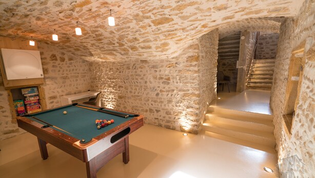 Una mesa de billar en una habitación con paredes de piedra y techo de madera. Se ve una escalera que conduce a otro nivel.