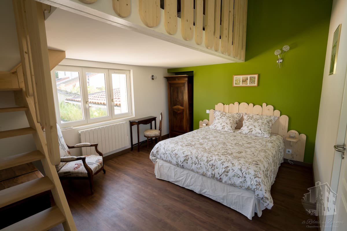 Un dormitorio con escaleras de madera, una cama con colcha estampada floral, una pared de acento verde y una ventana con vistas a follaje.