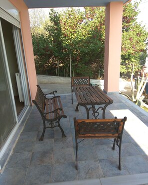 An outdoor patio with two wooden benches and a table, surrounded by a tiled floor and a covered area with a peach-colored wall.