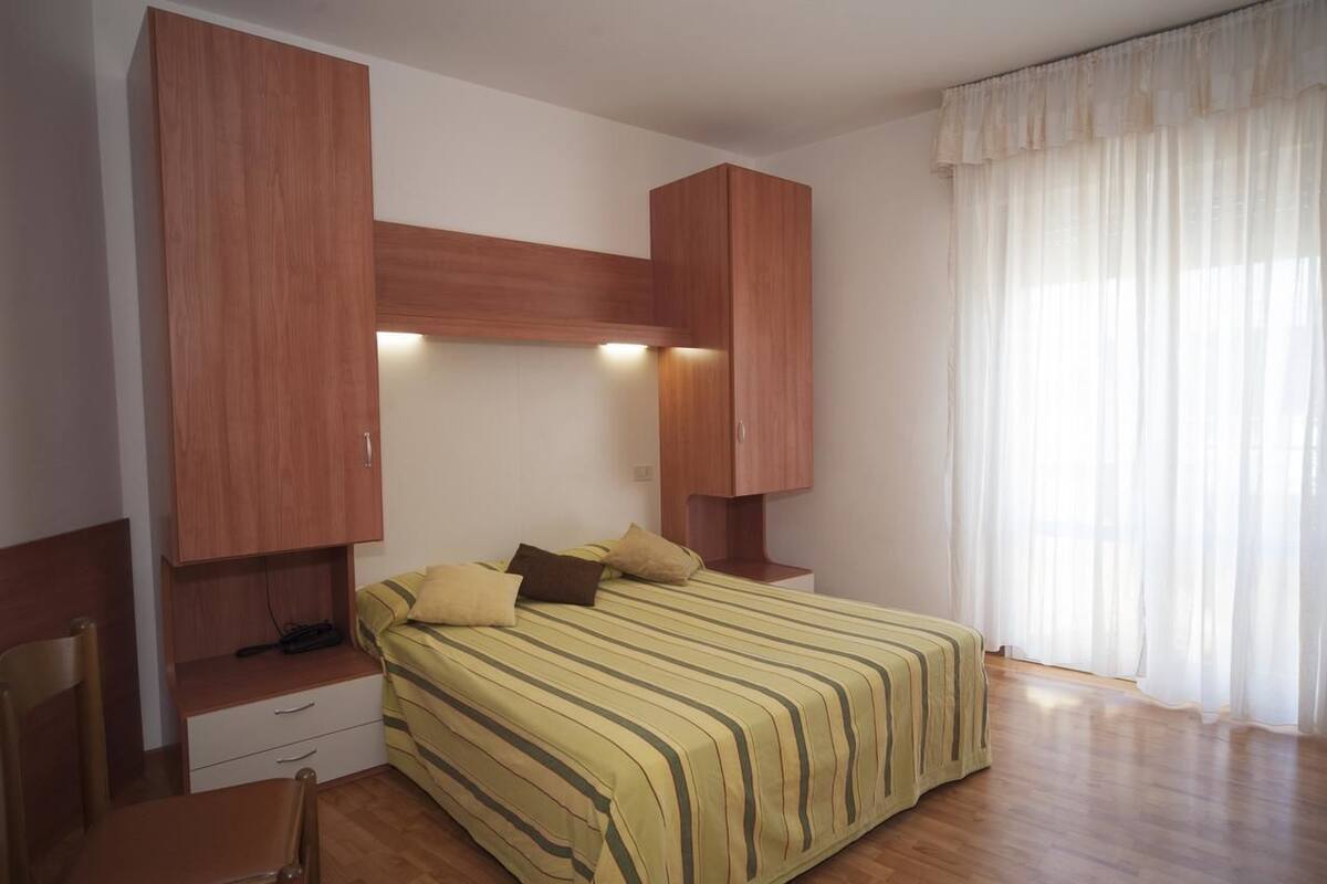 Un dormitorio con una cama, un escritorio y una silla. Hay armarios de madera sobre la cama y una ventana con cortinas.