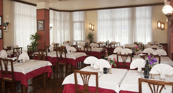 A restaurant with multiple tables set with white tablecloths and red table runners, wooden chairs, and wall-mounted light fixtures. - Hotel Galles (Jesolo)