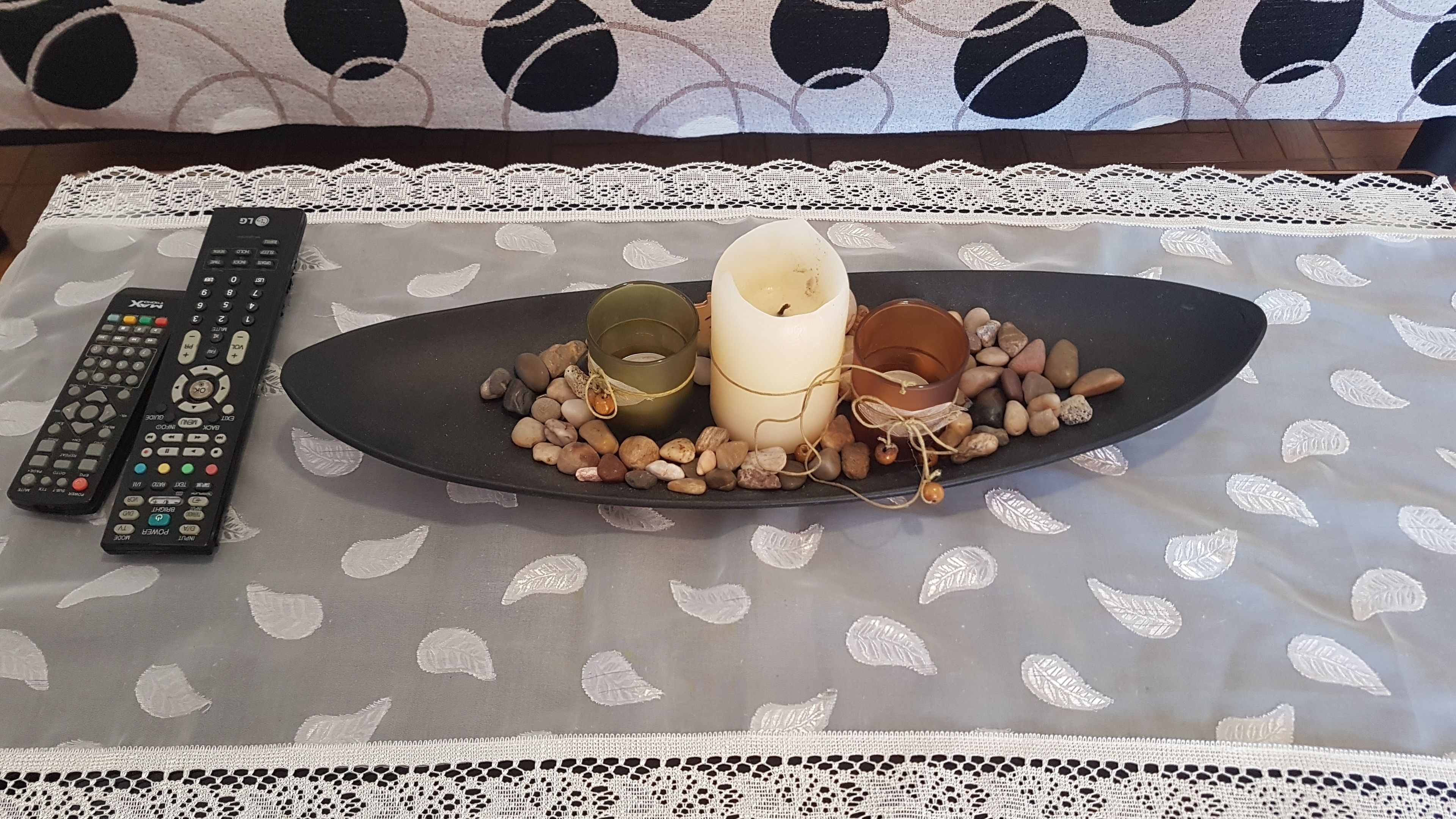 A table with a lace tablecloth, a candle, a plate with pebbles, and two remote controls.