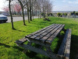 Una mesa de picnic en un parque con césped, árboles y una vista distante de una carretera y vehículos.