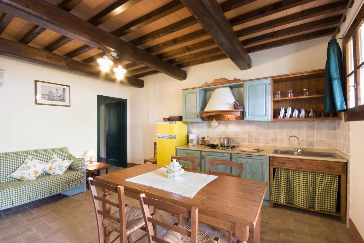 A cozy kitchen with wooden beams, a dining table with chairs, a green checkered sofa, and a small refrigerator.