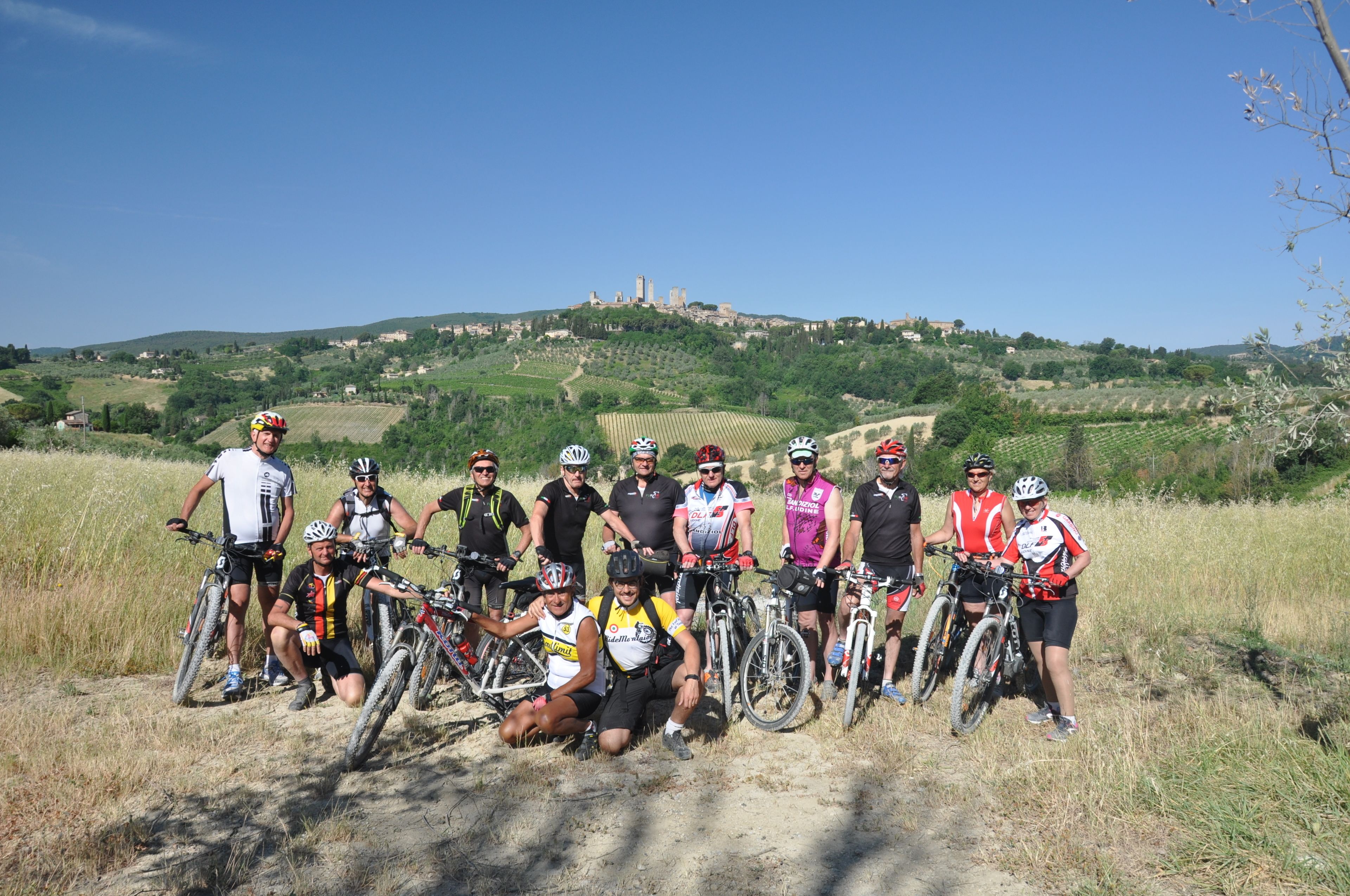 Un groupe de cyclistes pose sur un chemin de terre, avec en arrière-plan une vue panoramique sur des collines verdoyantes et un château.