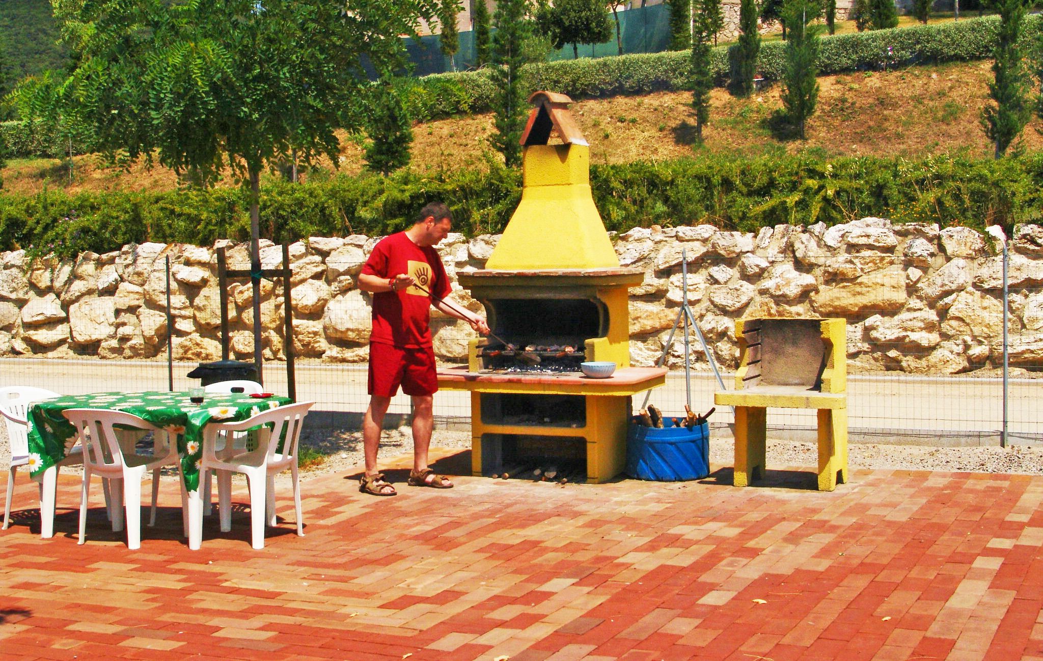 A man grilling outdoors with a yellow chimney, a table with chairs, and a stone wall in the background.