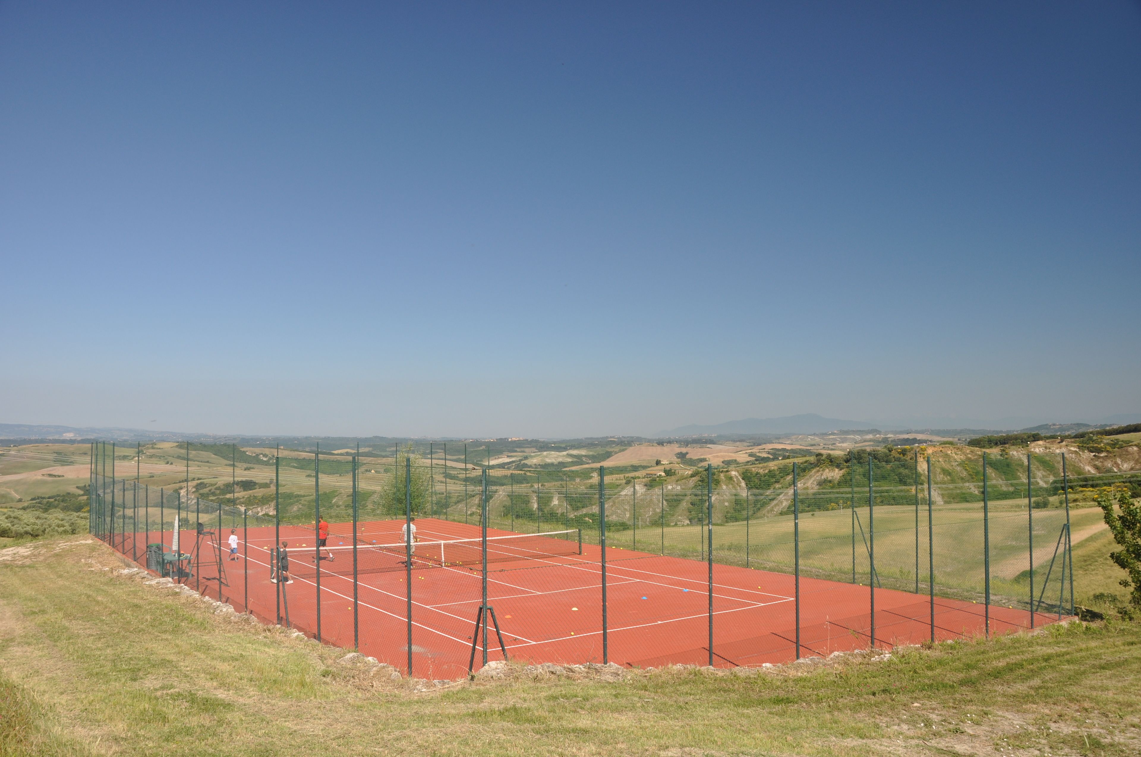 A tennis court with a net, surrounded by a green fence, set against a hilly landscape.