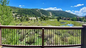 A scenic view from a balcony with a railing, overlooking a mountainous landscape with a river, buildings, and greenery. - Cinnamon Ridge B304 by SummitCove Vacation Lodging (Keystone)