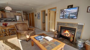 A living room with a fireplace, a television mounted above it, and a dining area with wooden furniture. - Cinnamon Ridge B304 by SummitCove Vacation Lodging (Keystone)