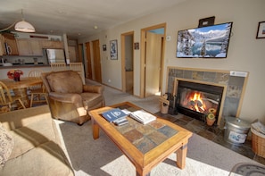 A living room with a fireplace, a television mounted above it, and a dining area with wooden furniture. - Cinnamon Ridge B304 by SummitCove Vacation Lodging (Keystone)
