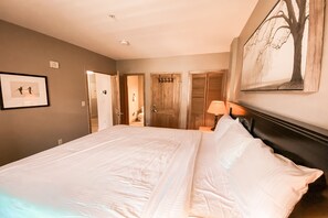 A large bed with white linens, a nightstand with a lamp, a framed picture on the wall, and a door leading to another room.