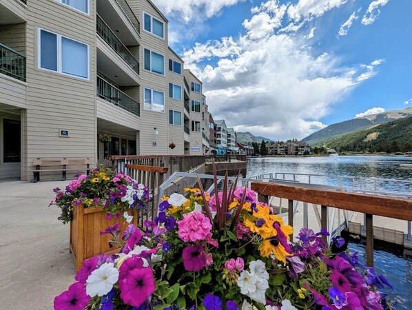 A waterfront hotel with balconies overlooking a lake, a wooden deck with flower arrangements, and a clear view of the mountains in the background.