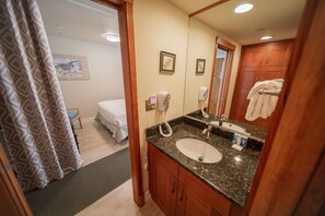 A bathroom with a sink, mirror, towel rack, and a bed in the adjacent room.