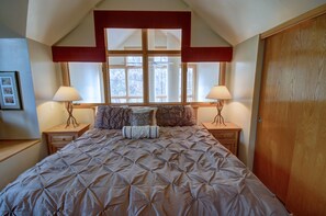 Quarto com uma cama grande, duas mesas de cabeceira com luminárias, um guarda-roupa de madeira e uma janela com vista para árvores.