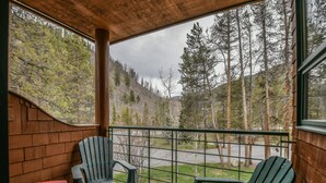 Une terrasse en bois avec deux fauteuils Adirondack verts et une petite table, donnant sur une zone boisée avec une route en arrière-plan.